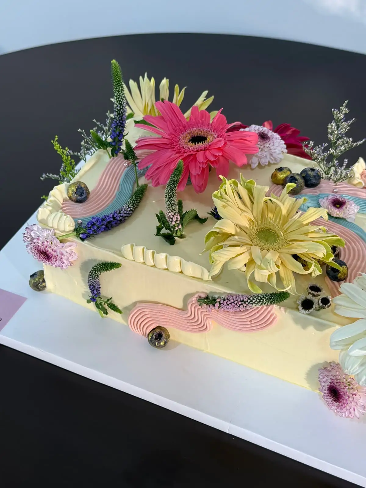 Pastel celebration cake decorated with fresh pink and yellow flowers, blueberries, and piped cream.
