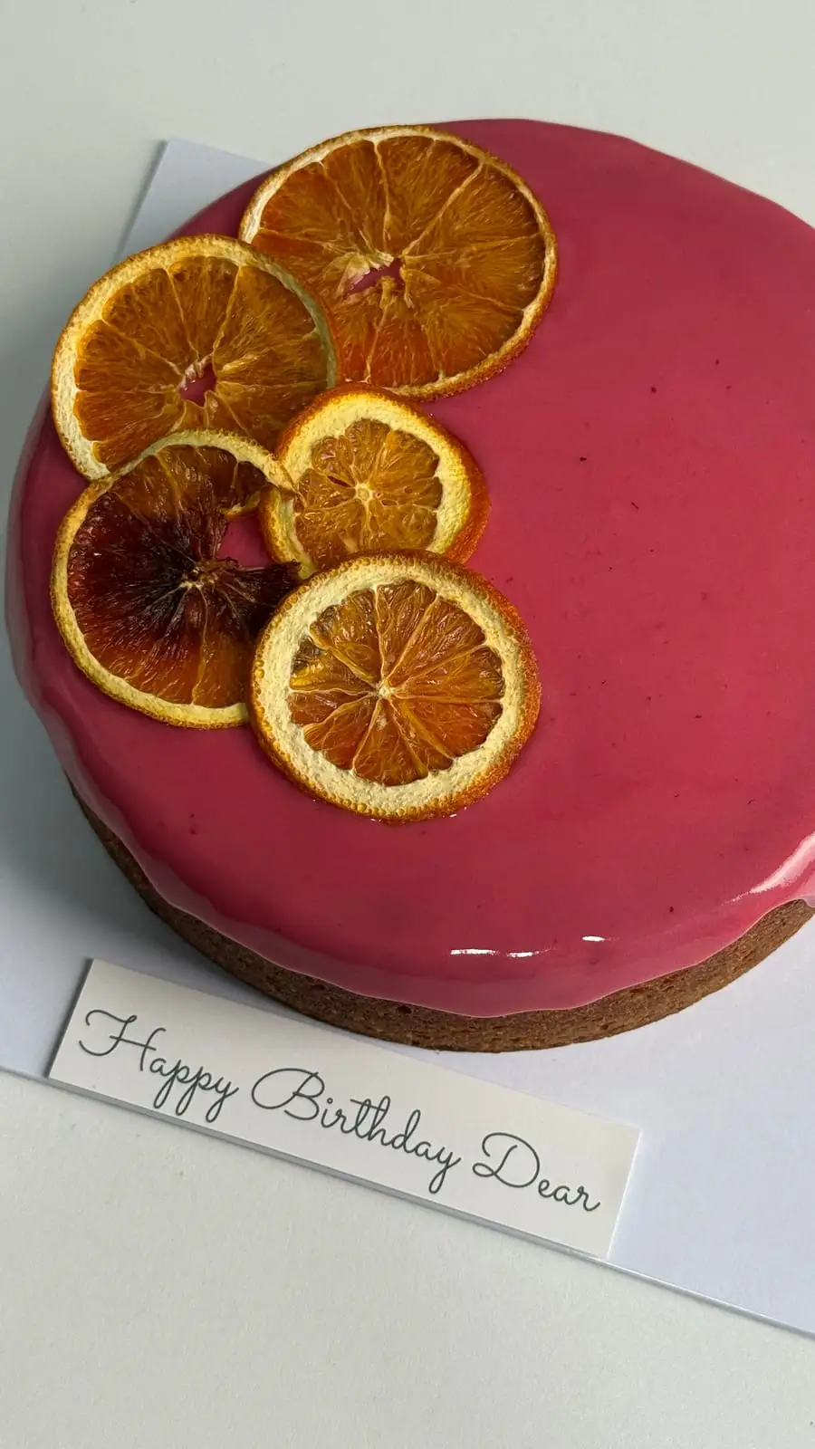 Glossy pink celebration cake decorated with dried orange slices and a handwritten birthday message.